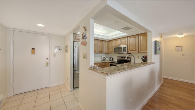 kitchen featuring stainless steel appliances, light stone counters, a tray ceiling, kitchen peninsula, and light brown cabinets