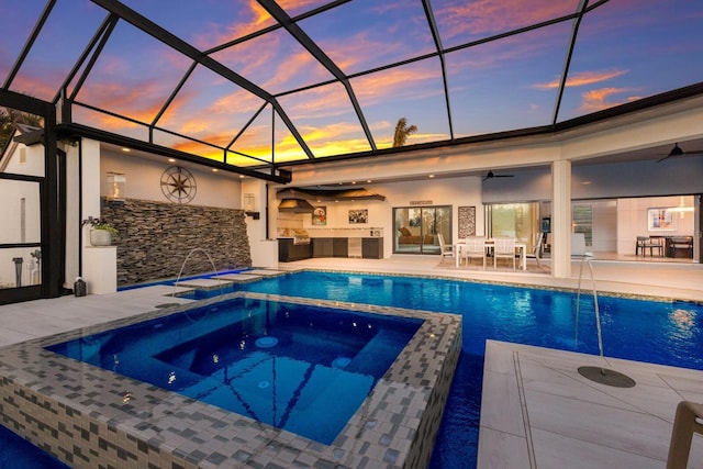 pool at dusk featuring a patio, exterior kitchen, an in ground hot tub, a lanai, and pool water feature