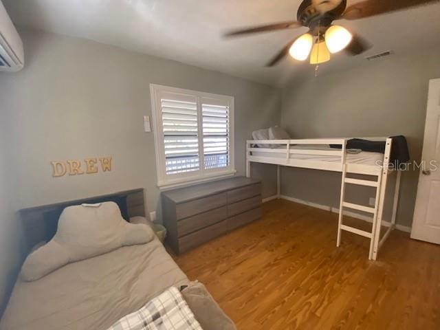 bedroom with a wall mounted AC, wood finished floors, visible vents, and a ceiling fan