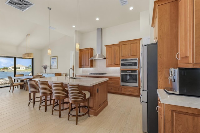 kitchen with a breakfast bar area, appliances with stainless steel finishes, hanging light fixtures, an island with sink, and wall chimney exhaust hood