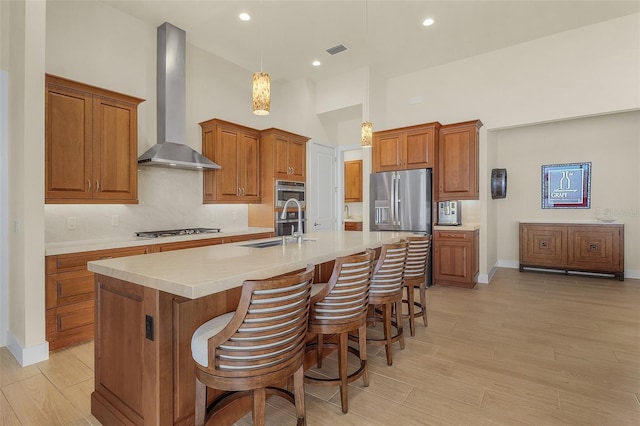 kitchen featuring sink, hanging light fixtures, stainless steel appliances, a kitchen island with sink, and wall chimney range hood