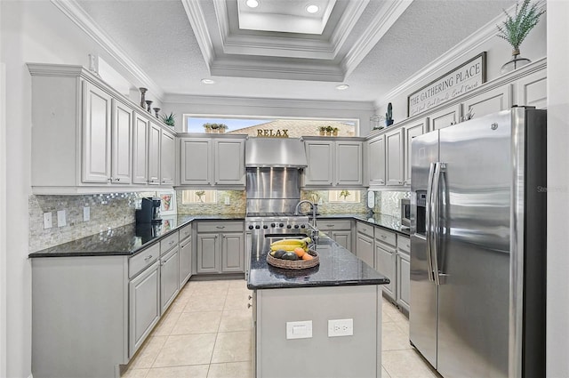 kitchen featuring stainless steel fridge with ice dispenser, a tray ceiling, dark stone counters, a kitchen island with sink, and wall chimney range hood