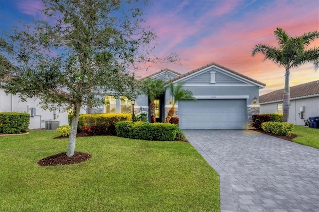 single story home featuring central AC unit, a lawn, stone siding, an attached garage, and decorative driveway