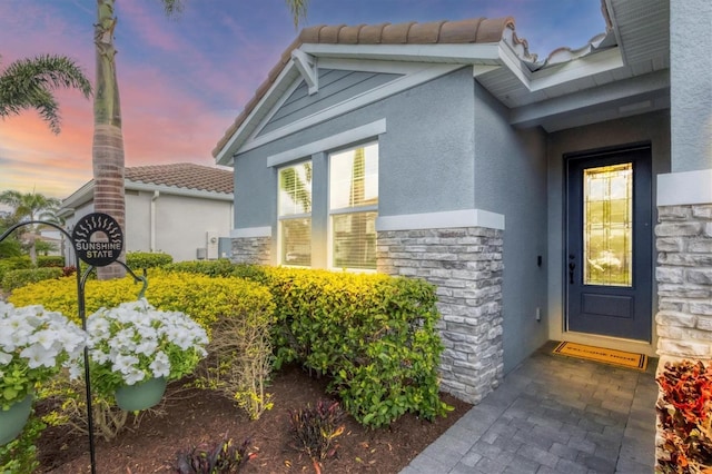 exterior entry at dusk with stone siding, a tiled roof, and stucco siding