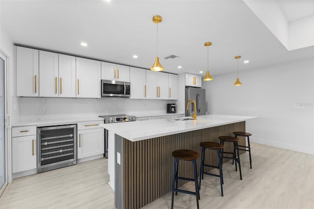 kitchen featuring white cabinetry, appliances with stainless steel finishes, an island with sink, pendant lighting, and beverage cooler