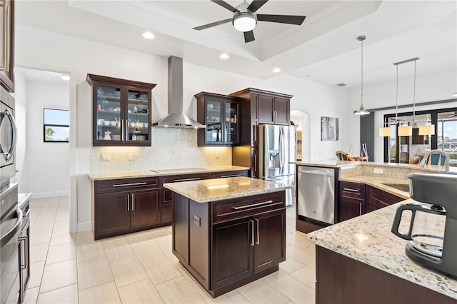 kitchen featuring appliances with stainless steel finishes, a center island, decorative light fixtures, a raised ceiling, and wall chimney exhaust hood