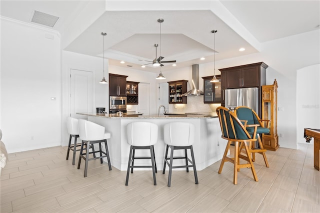 kitchen featuring wall chimney exhaust hood, hanging light fixtures, stainless steel microwave, light stone countertops, and backsplash