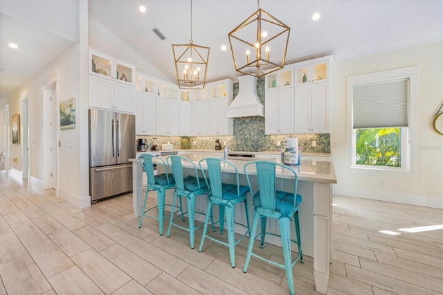 kitchen featuring stainless steel fridge, premium range hood, white cabinets, a center island with sink, and decorative light fixtures