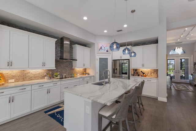 kitchen with sink, white cabinets, wall chimney range hood, stainless steel refrigerator with ice dispenser, and a center island with sink