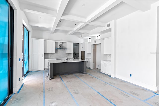 kitchen with wall chimney range hood, sink, coffered ceiling, white cabinets, and a center island with sink