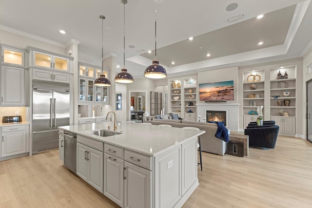 kitchen with sink, appliances with stainless steel finishes, a kitchen island with sink, light stone countertops, and a raised ceiling