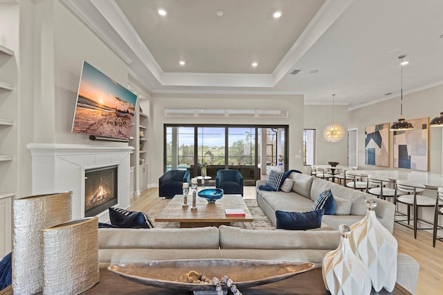 living room with a tray ceiling, built in shelves, ornamental molding, and light wood-type flooring