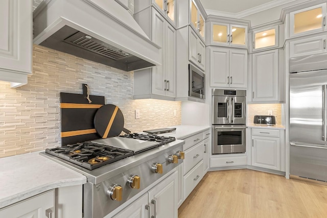 kitchen featuring custom exhaust hood, white cabinetry, built in appliances, light wood-type flooring, and backsplash