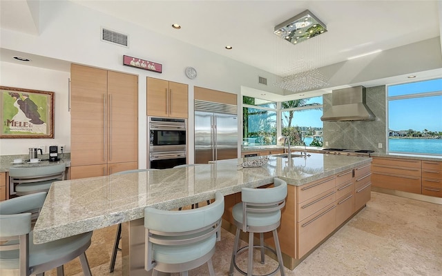 kitchen with wall chimney range hood, stainless steel appliances, a kitchen breakfast bar, a water view, and light stone counters