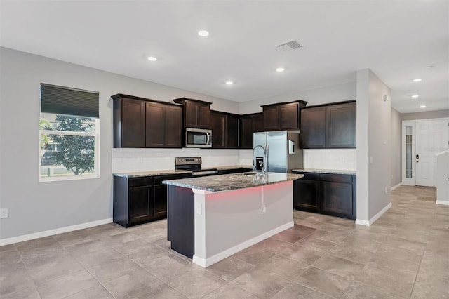 kitchen featuring dark brown cabinetry, appliances with stainless steel finishes, light stone countertops, a kitchen island with sink, and decorative backsplash