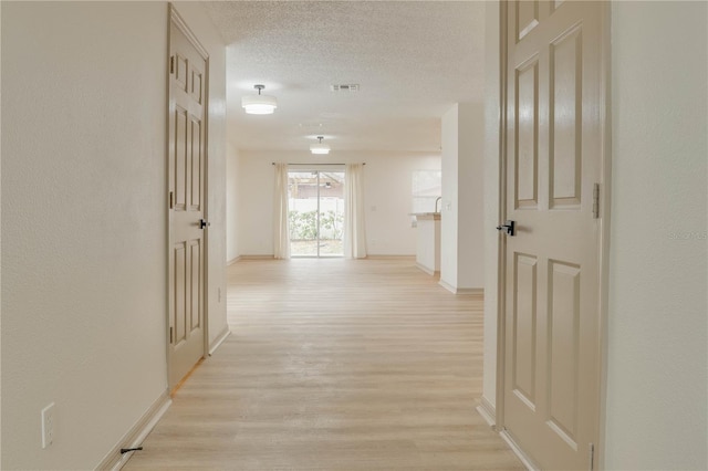 hallway with a textured ceiling, light wood-type flooring, visible vents, and baseboards