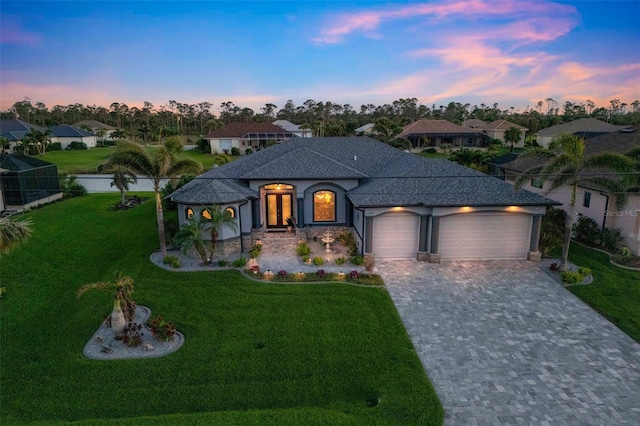 french country home with decorative driveway, roof with shingles, a lawn, an attached garage, and a residential view