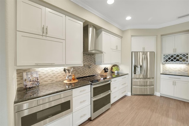 kitchen featuring dark stone counters, appliances with stainless steel finishes, ornamental molding, wall chimney range hood, and white cabinetry