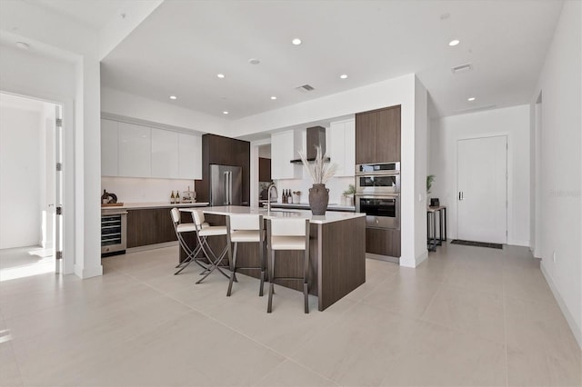 kitchen featuring wine cooler, wall chimney exhaust hood, a breakfast bar area, white cabinetry, and a kitchen island with sink