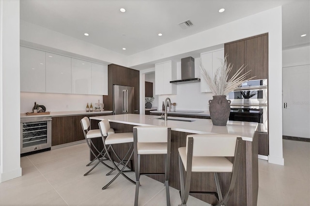 kitchen featuring appliances with stainless steel finishes, a breakfast bar, wine cooler, a kitchen island with sink, and wall chimney exhaust hood