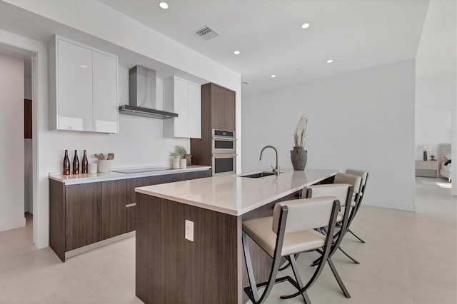 kitchen with dark brown cabinetry, wall chimney exhaust hood, sink, white cabinetry, and a center island with sink