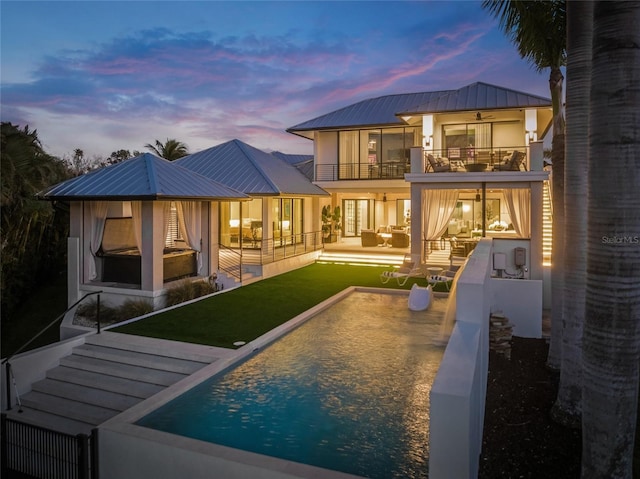 back house at dusk featuring a patio area, a balcony, a yard, outdoor lounge area, and ceiling fan