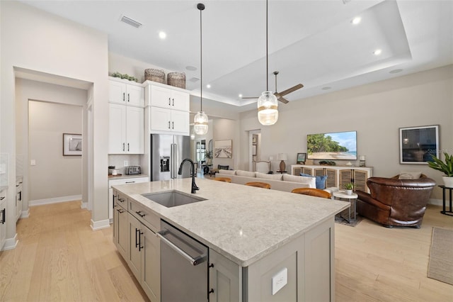 kitchen with light wood finished floors, a center island with sink, appliances with stainless steel finishes, a tray ceiling, and a sink