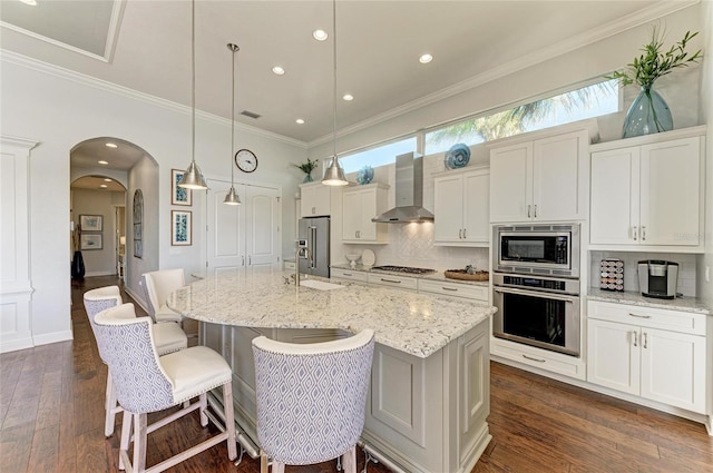 kitchen featuring stainless steel appliances, a kitchen island with sink, decorative light fixtures, and wall chimney exhaust hood