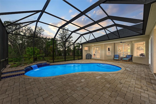 pool at dusk featuring pool water feature, ceiling fan, a patio, and glass enclosure