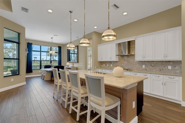 kitchen with wall chimney range hood, an island with sink, decorative light fixtures, and white cabinets