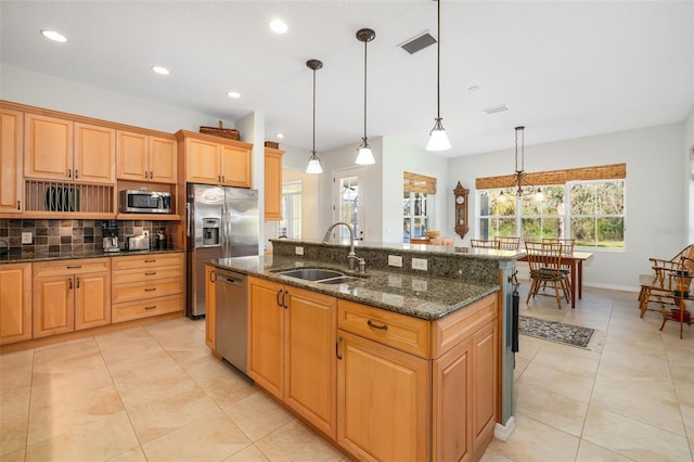 kitchen featuring sink, stainless steel appliances, a center island with sink, decorative light fixtures, and dark stone counters