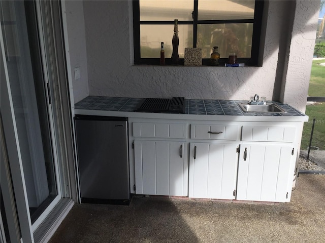 kitchen with tile countertops, a textured wall, a sink, white cabinets, and freestanding refrigerator