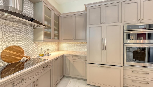 kitchen featuring marble finish floor, glass insert cabinets, stainless steel double oven, light stone countertops, and wall chimney exhaust hood