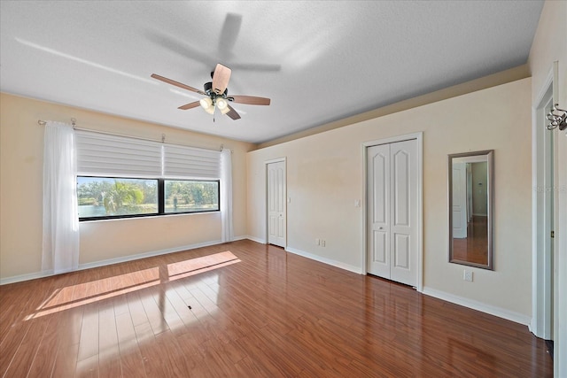 unfurnished bedroom with a textured ceiling, ceiling fan, wood finished floors, baseboards, and two closets