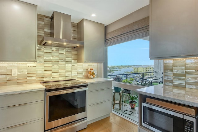 kitchen with light stone counters, stainless steel appliances, wall chimney range hood, light wood-type flooring, and backsplash