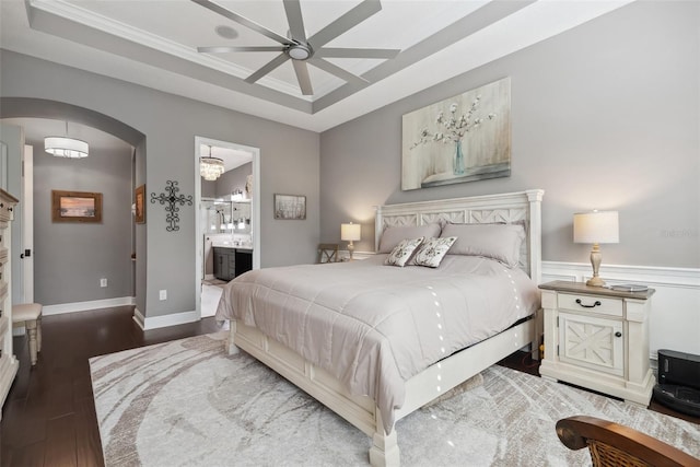 bedroom featuring arched walkways, wood finished floors, baseboards, a tray ceiling, and ensuite bath