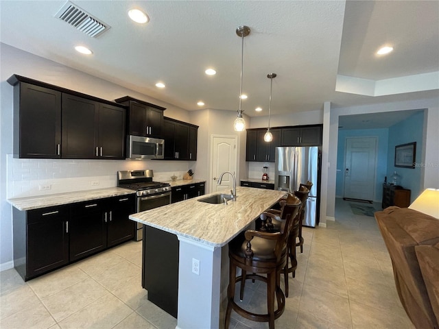 kitchen with a center island with sink, sink, decorative light fixtures, stainless steel appliances, and light stone countertops