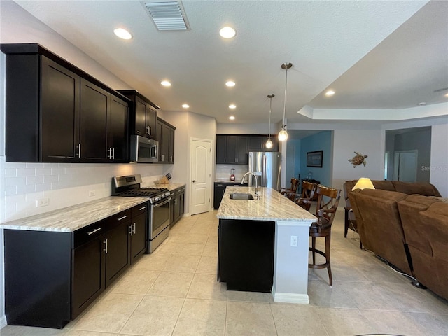 kitchen with backsplash, decorative light fixtures, stainless steel appliances, an island with sink, and light stone countertops