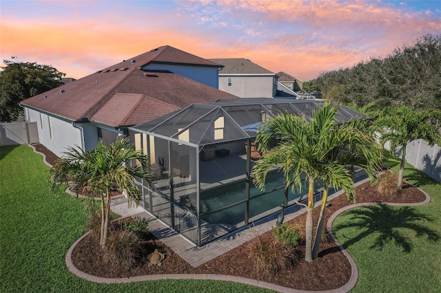 back of house at dusk featuring a patio, a lanai, fence, a lawn, and stucco siding