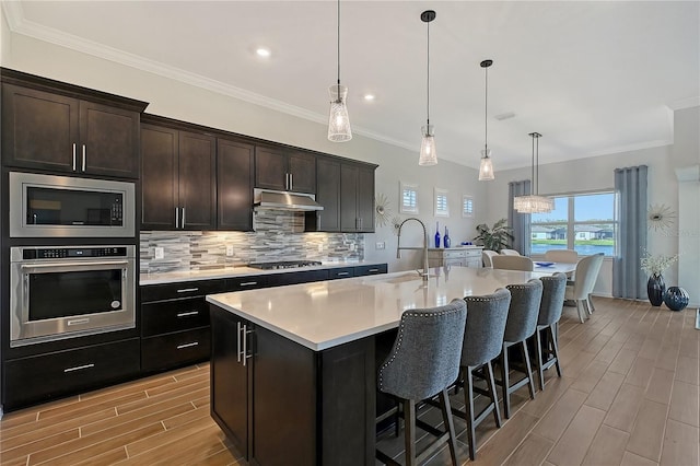 kitchen with a center island with sink, light countertops, hanging light fixtures, appliances with stainless steel finishes, and under cabinet range hood