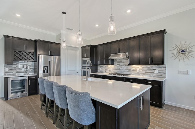 kitchen featuring wine cooler, a kitchen island with sink, under cabinet range hood, light countertops, and appliances with stainless steel finishes