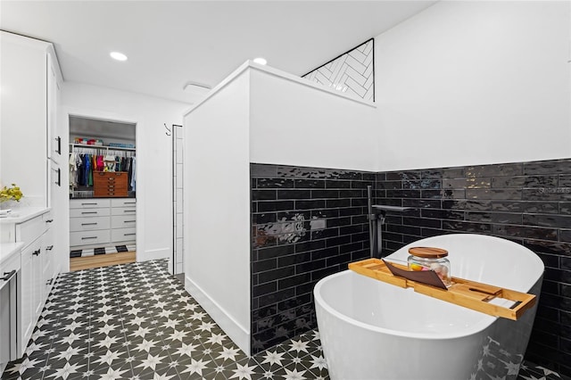 full bathroom featuring a walk in closet, tile walls, recessed lighting, vanity, and a freestanding tub