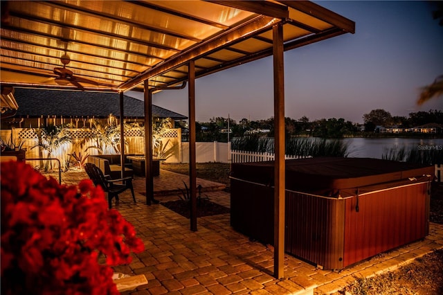 patio terrace at dusk with a water view, a hot tub, and fence