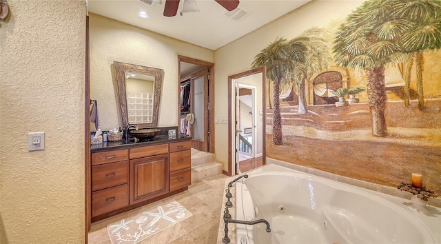 full bath featuring a walk in closet, a textured wall, ceiling fan, vanity, and a tub with jets