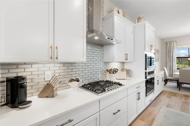 kitchen with appliances with stainless steel finishes, wall chimney range hood, light hardwood / wood-style floors, backsplash, and white cabinetry