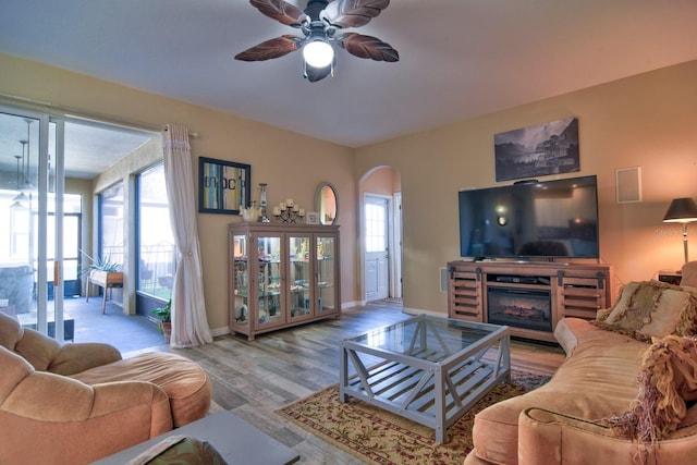 living room featuring arched walkways, wood finished floors, a ceiling fan, baseboards, and a glass covered fireplace