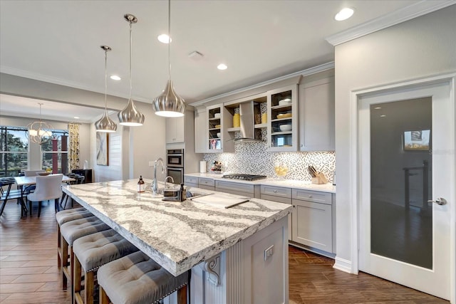 kitchen with light stone counters, wall chimney range hood, an island with sink, glass insert cabinets, and pendant lighting