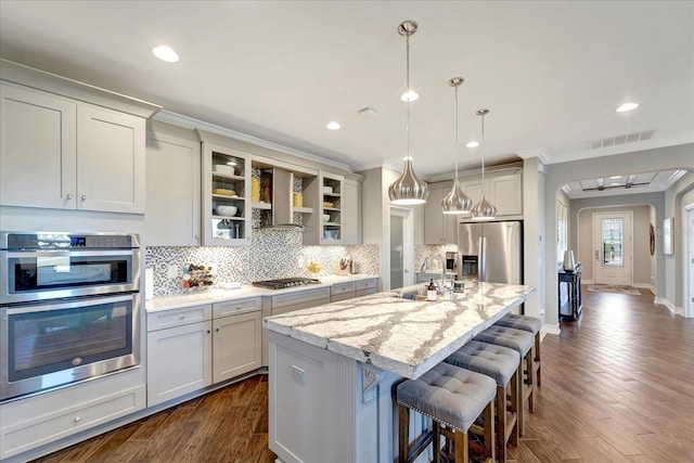 kitchen with glass insert cabinets, light stone counters, a kitchen island with sink, stainless steel appliances, and wall chimney range hood