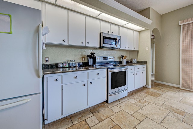 kitchen featuring arched walkways, stone tile floors, stainless steel appliances, white cabinetry, and baseboards