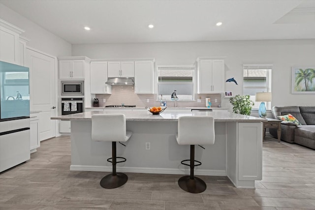 kitchen featuring stainless steel appliances, white cabinetry, a kitchen island, under cabinet range hood, and a kitchen bar